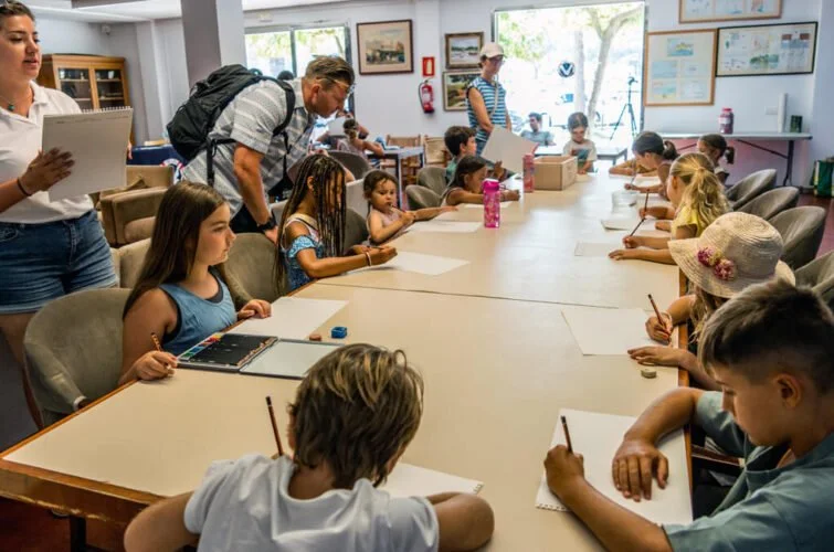 Niños y niñas en una clase de arte, pintando y dibujando en una mesa grande, con adultos supervisando en un aula luminosa.