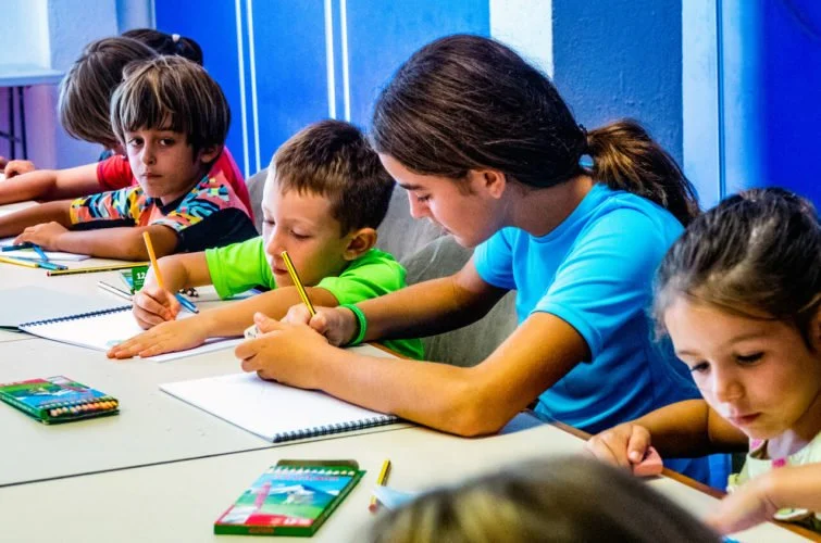 Una maestra enseñando a un grupo de niños en un aula, todos haciendo dibujo con lápices.
