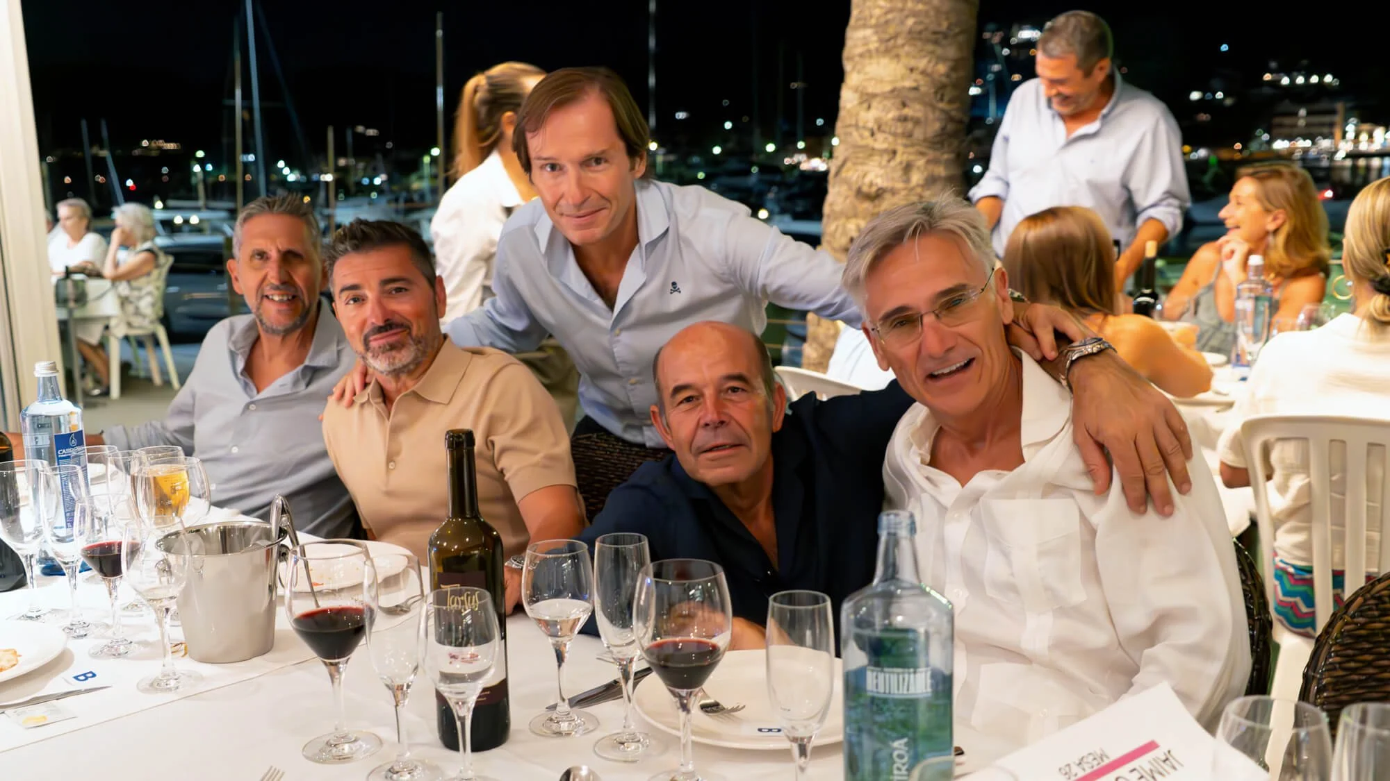 Grupo de personas en una cena al aire libre en la noche, algunos brindando, con vino y bebidas en la mesa, en un restaurante cerca del puerto con yates y luces en el fondo.