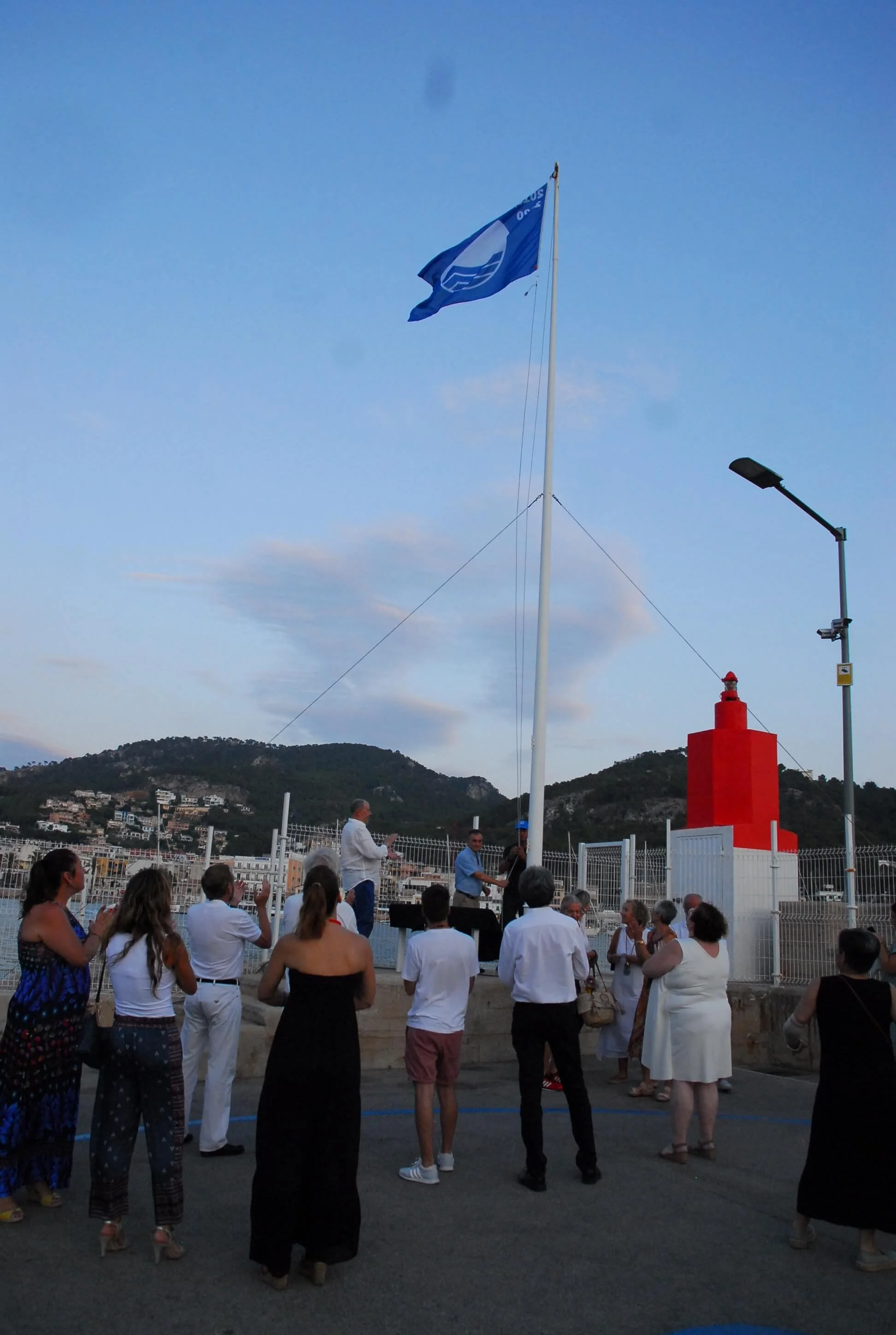 Reunión de personas en una plaza con una bandera azul en un mástil y un faro rojo, durante el atardecer, con edificios y montañas de fondo.