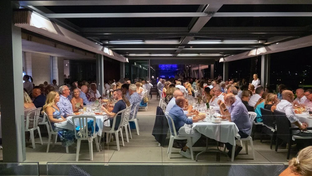Cena de una cena social en un espacio cerrado, con muchas mesas y personas comiendo y conversando, ambiente formal y elegante