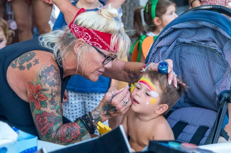 Una mujer con tatuajes y gafas pinta la cara a un niño en un evento al aire libre, otros niños y adultos están presentes en el fondo.