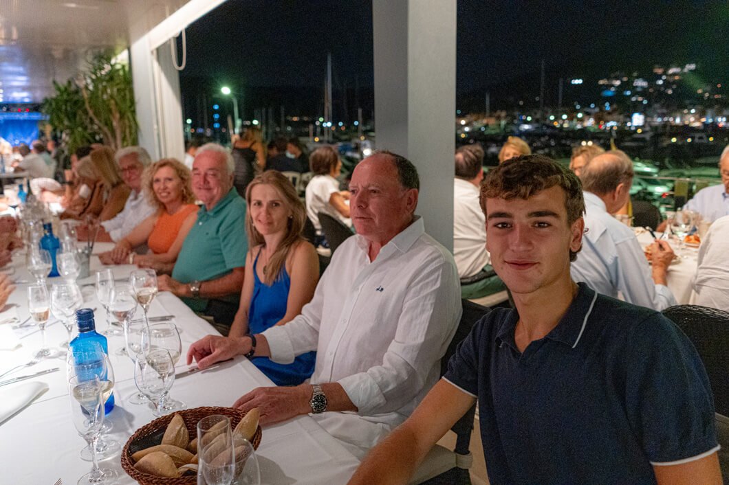 Grupo de personas en una cena en un restaurante, algunas sonriendo, en una mesa con copas y botellas, con fondo de luces nocturnas y un área exterior visible.