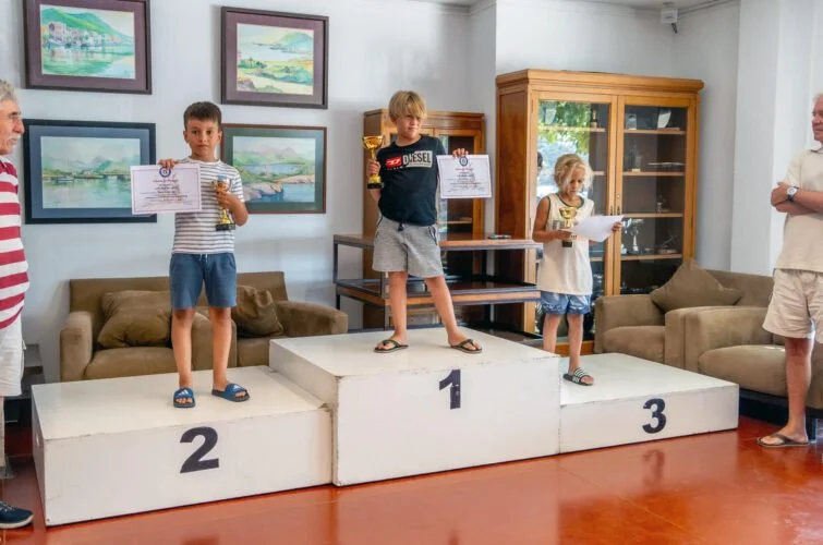 Niños en un podio ganando una competencia, con adultos a los lados en una sala con cuadros y armario de madera.