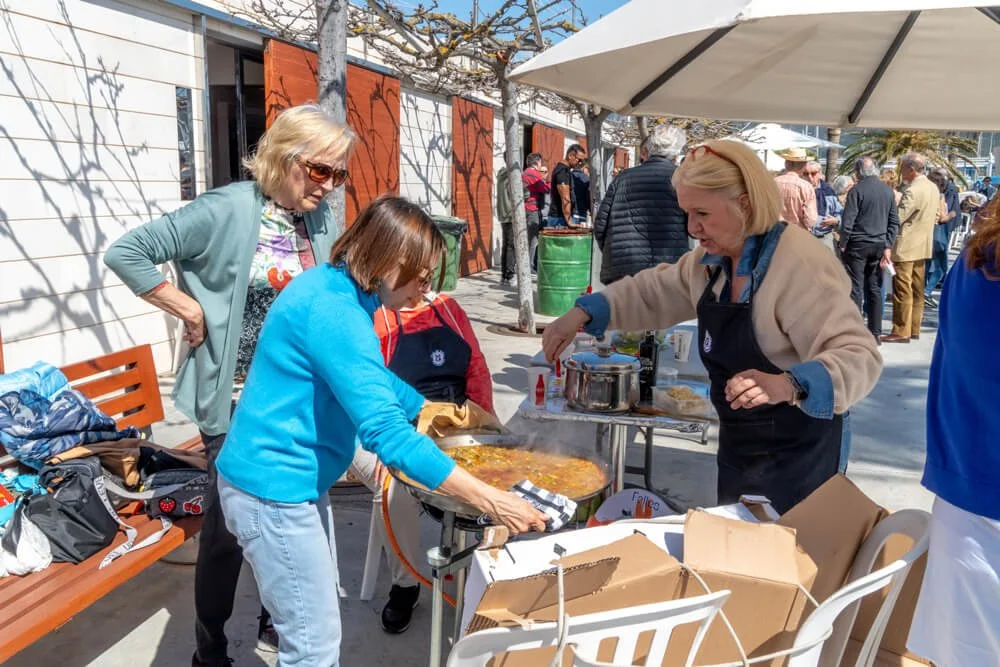Varias personas están en un puesto de comida al aire libre, sirviendo y comiendo comida en platos y utensilios desechables, con un ambiente social y concurrido.