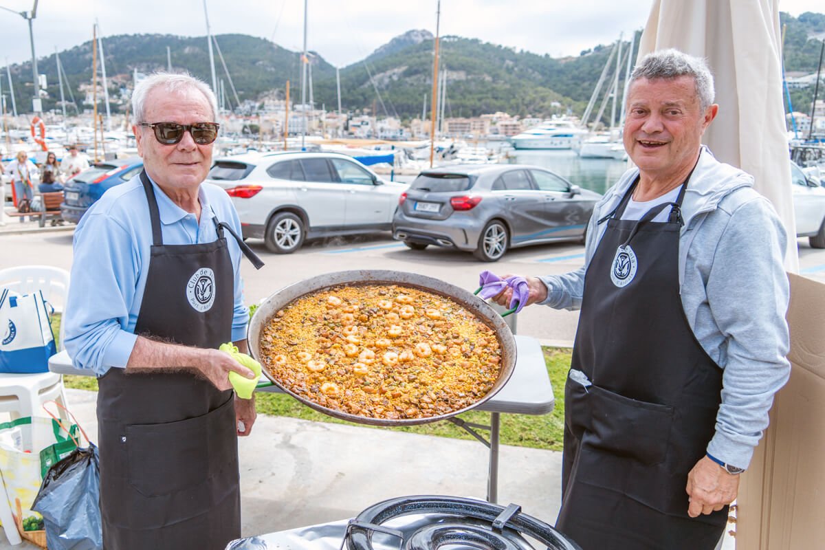 Dos hombres cocinando paella en un evento frente al puerto con montañas al fondo.