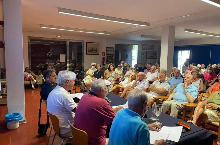 Reunión de un grupo de personas en un salón, algunas sentadas en sillas y otras en una mesa en la parte frontal, participando en una conferencia o discusión.