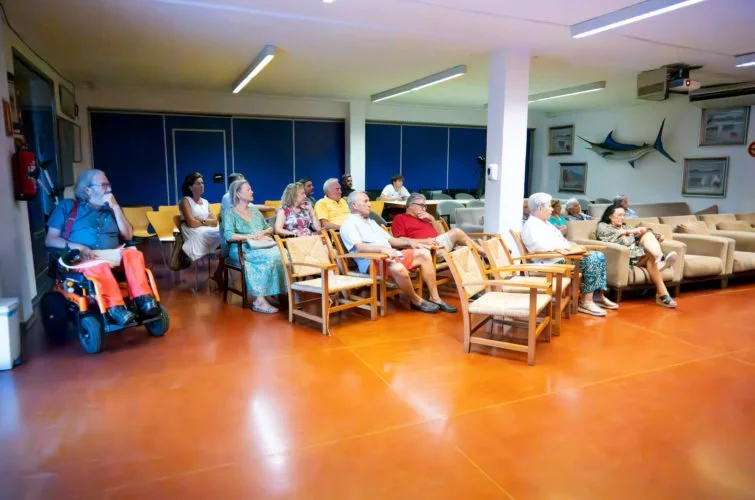 Grupo de personas sentadas en sillas y sofás en una sala, algunos son adultos mayores, en un entorno que parece ser un centro de atención o sala de espera, decorada con tiburón de madera en la pared y cuadros.