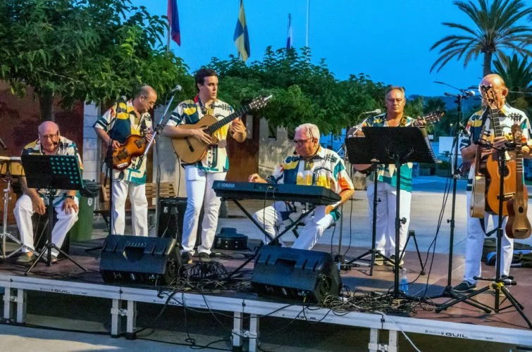 Grupo de músicos tocando en un escenario al aire libre, con árboles y banderas de fondo, usando camisetas con colores vivos.