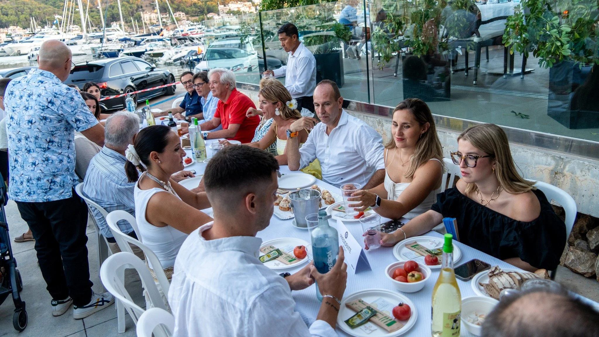 Un grupo de personas sentadas en una mesa al aire libre en un evento, con una vista de un puerto con barcos y coches de fondo, mientras un hombre con camiseta de flores sirve comida.