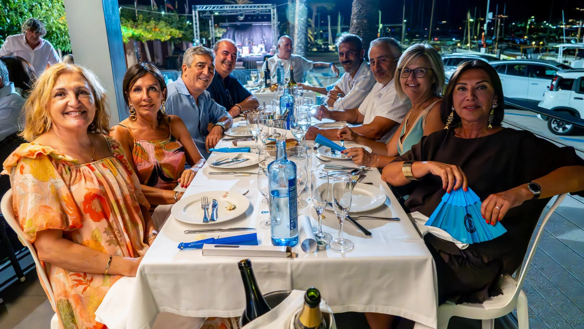 Grupo de personas cenando en un restaurante al aire libre durante la noche, con vista a un puerto con botes y luces en el fondo.