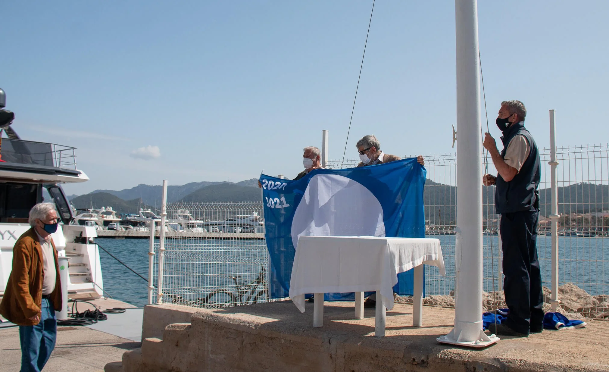 Personas en un muelle con banderas y equipo, durante un evento en el puerto, con montañas y barcos al fondo en un día soleado.