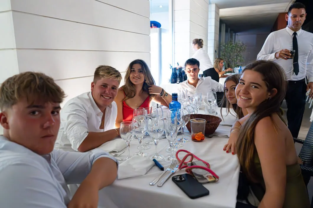 Grupo de jóvenes en una mesa de restaurante, sonriendo y disfrutando una comida elegante con copas y vajilla fina, en un ambiente formal y bien iluminado.
