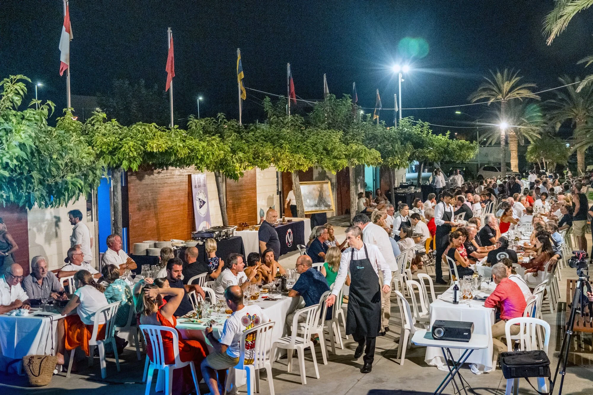 Una celebración al aire libre por la noche con muchas personas sentadas en mesas, algunos camareros atendiendo, banderas en el fondo, palmeras y árboles y luces en el cielo.