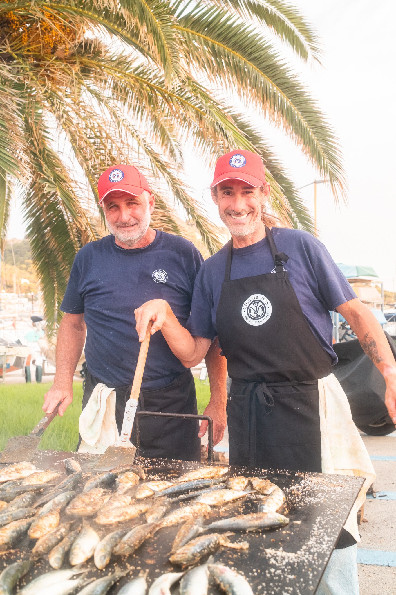 Dos hombres preparando pescado a la parrilla en un lugar al aire libre, con palmeras de fondo y todos llevan camisetas y gorras rojas con un logotipo, uno de ellos sonriente.