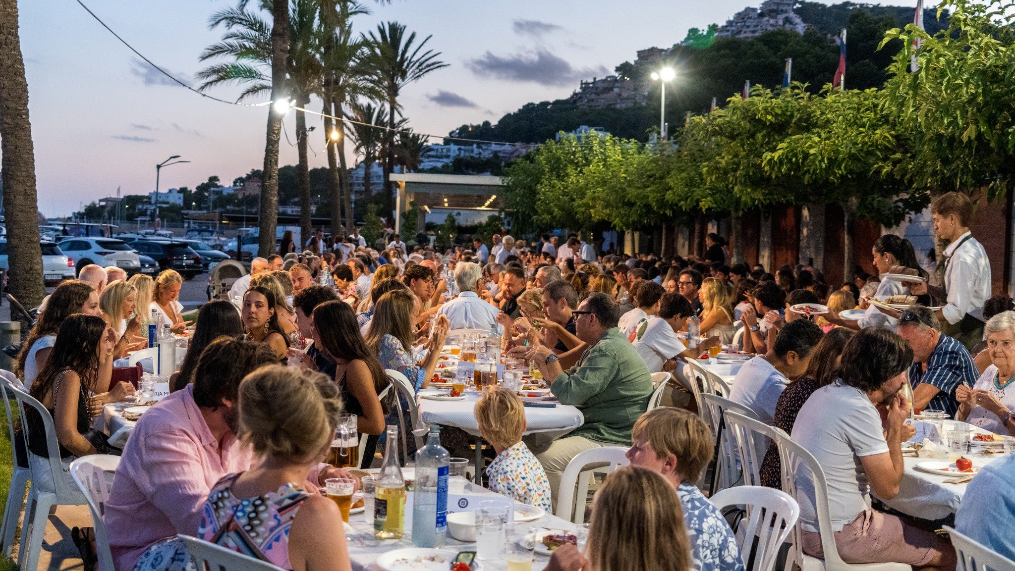 Una multitud de personas cena al aire libre en una terraza junto al mar, con palmeras y una colina con edificios y banderas de fondo, durante el atardecer.