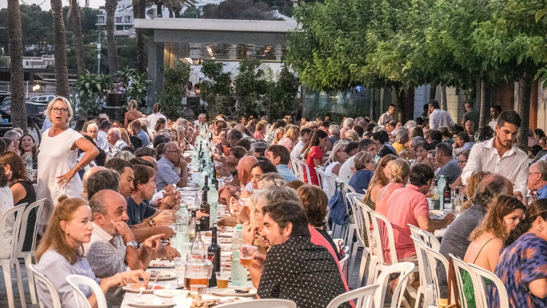 Una gran reunión al aire libre con muchas personas sentadas en mesas largas, disfrutando de una cena en un ambiente nocturno con árboles y un edificio en el fondo.