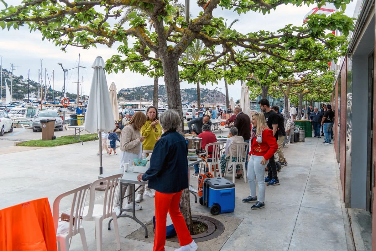 Personas disfrutando en una terraza junto al mar con árboles, mesas, sillas y yates en el muelle en un día nublado.