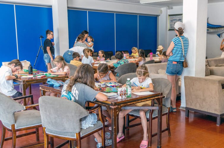 Niños y niñas en una clase artísticas, algunos dibujos y materiales sobre las mesas, en un salón con colores vivos y gente supervisando