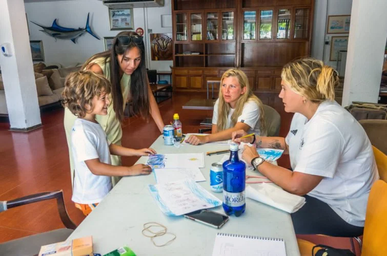 Cuatro personas en una reunión en un ambiente interior, con dos mujeres y dos niños, uno de los niños está mostrando su trabajo. Hay botellas de agua y papeles sobre la mesa.