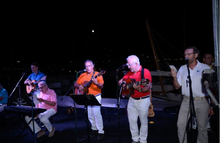 Grupo de músicos tocando en un escenario al aire libre en la noche, con instrumentos como guitarra, teclados y micrófonos, en un ambiente de concierto frente a un muelle o puerto.