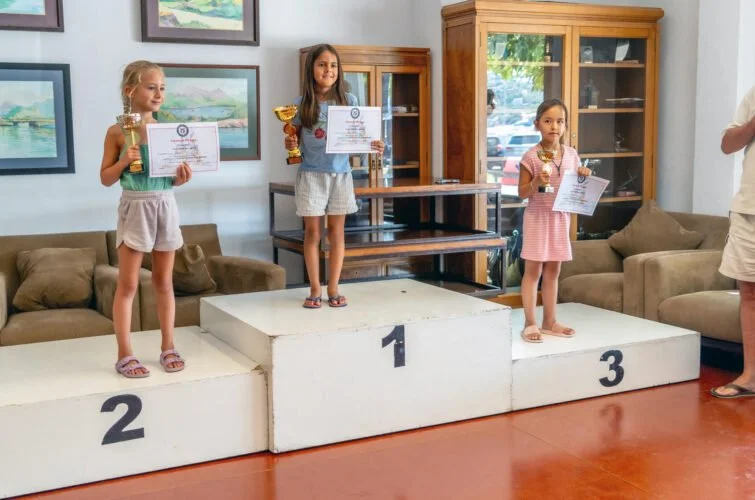 Tres niñas en un podio de premios, en el primer lugar, reciben diploma y trofeo, en segundo lugar, otra niña recibe diploma y trofeo, y en tercer lugar, una niña con diploma y trofeo. La escena parece ser una ceremonia de premiación infantil.