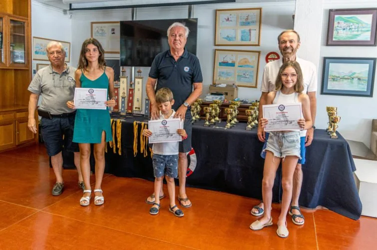 Grupo de seis personas, incluyendo niños y adultos, en una ceremonia de premiación con certificados y trofeos en una sala decorada con cuadros y un armario de madera.