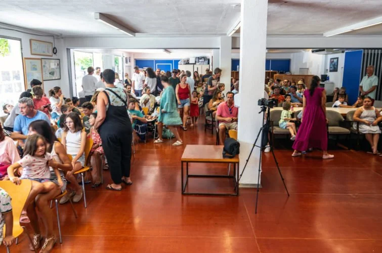 Sala llena de personas, niños y adultos, en lo que parece ser una espera o evento escolar, en un espacio con ventanas y sillas.