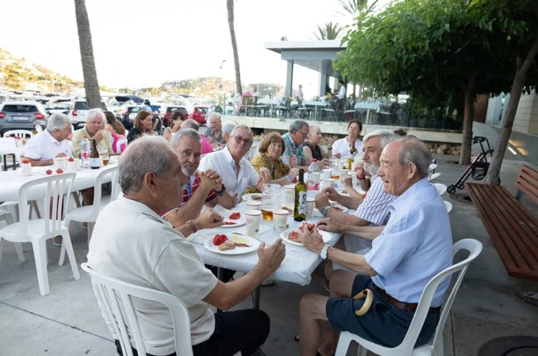 Grupo de personas mayores disfrutando de una comida al aire libre en un restaurante, con platos, bebidas y en un ambiente con árboles y vista al mar.