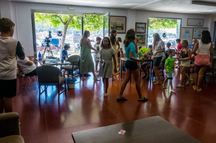 Niños y adultos en una sala, participando en una actividad social, con ventanas grandes que muestran árboles y barcos en el agua.