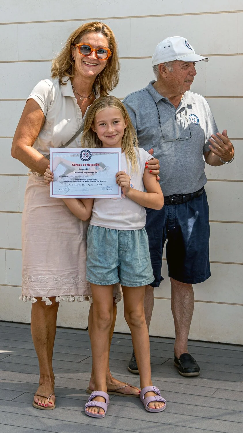 Una niña sostiene un certificado de participación en un curso de natación, acompañada por una mujer mayor y un hombre en un evento de natación. La mujer usa gafas naranjas y una blusa blanca, el hombre lleva una gorra blanca y camiseta gris, y la niñ