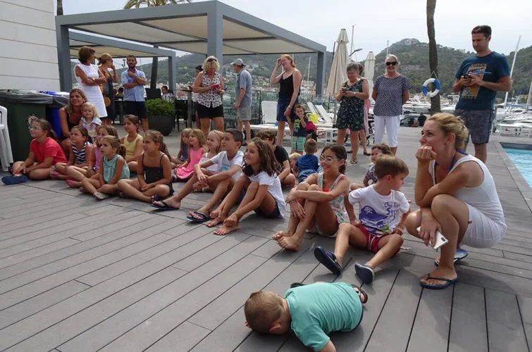 Grupo de niños y adultos en un muelle junto a yates, algunos niños sentados en el suelo y adultos de pie observando, ambiente de día soleado.