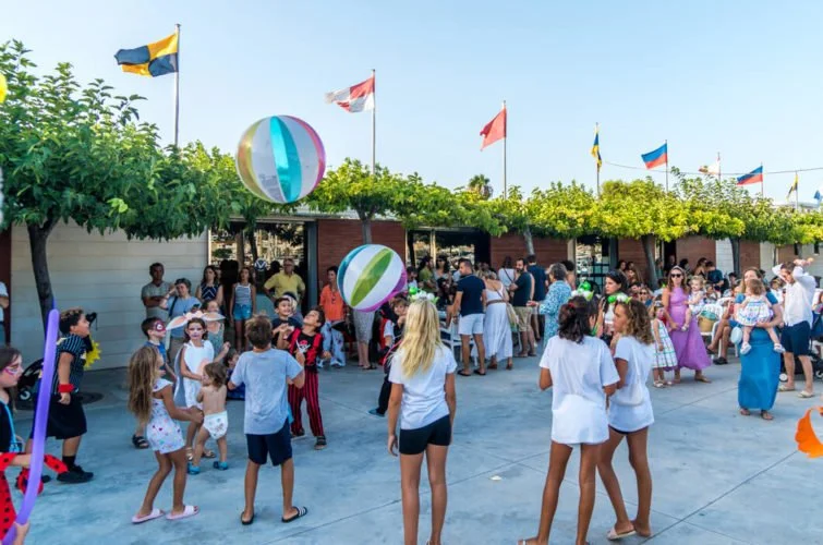 Personas y niños en una fiesta al aire libre con banderas, globos y decoraciones en un lugar con árboles y cielo despejado.