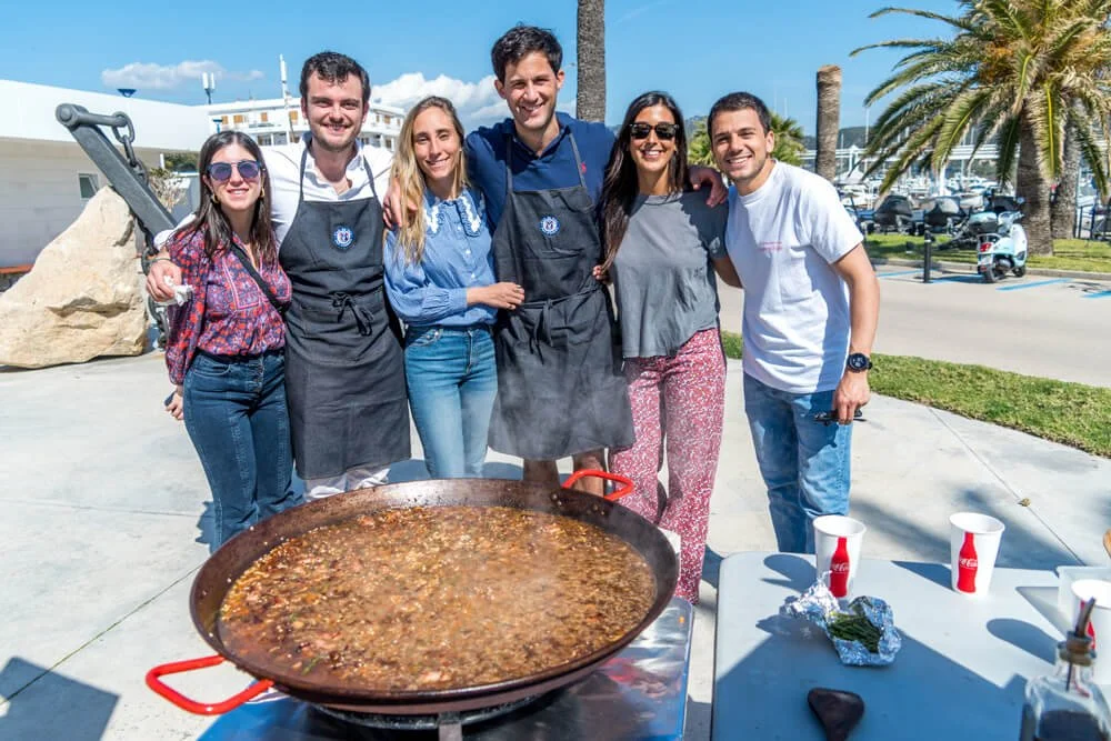 Grupo de seis personas sonriendo y posando para la foto al lado de una paella gigante en un ambiente al aire libre, con palmeras, un cielo brillante y mesas con vasos en primer plano.
