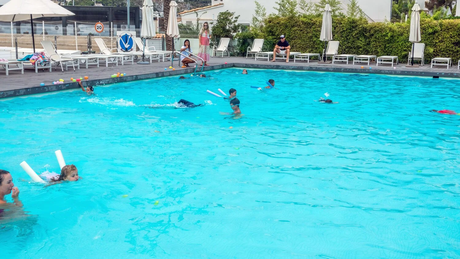 Niños y adultos disfrutando en una piscina, algunos niños con flotadores, rodeada de tumbonas, sombrillas y arbustos.