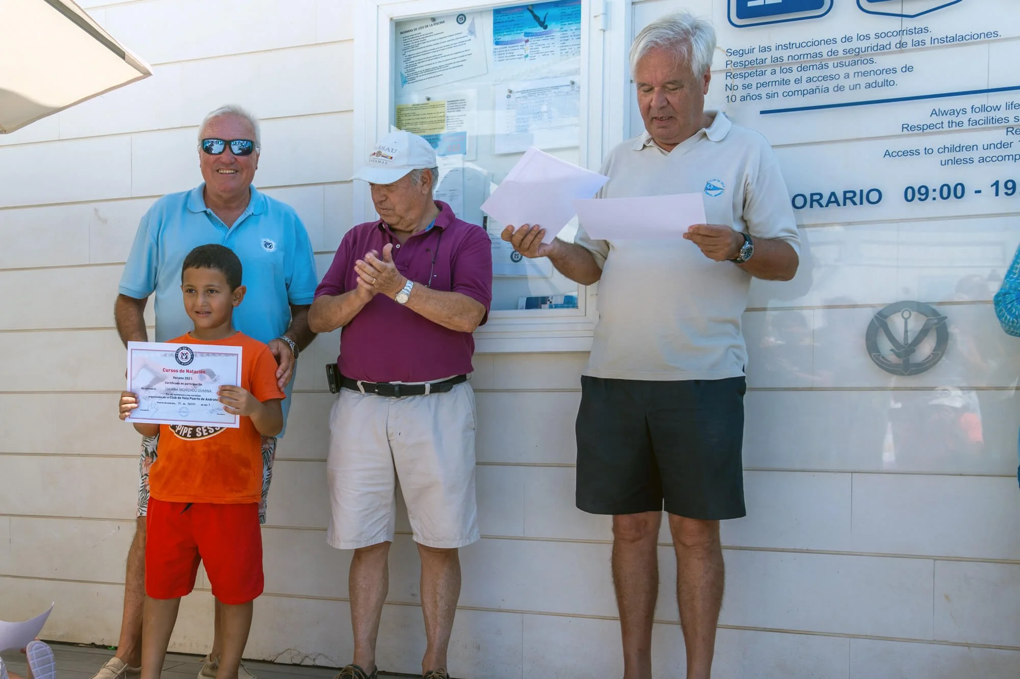 Niño recibiendo certificado en ceremonia de natación junto a adultos en un muro con información en español.