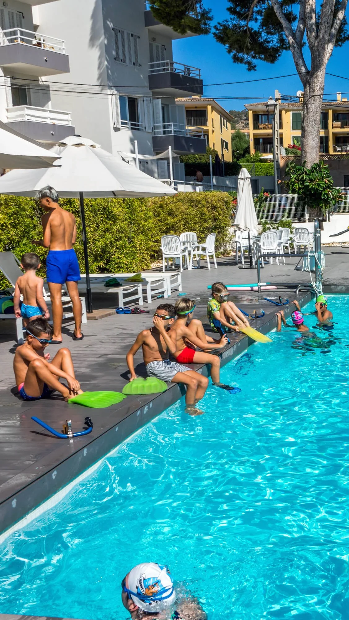 Niños y un adulto en la piscina, algunos niños se están preparando para nadar, otros están sentados en el borde y un adulto de pie junto a la piscina en un día soleado con edificios residenciales de fondo.