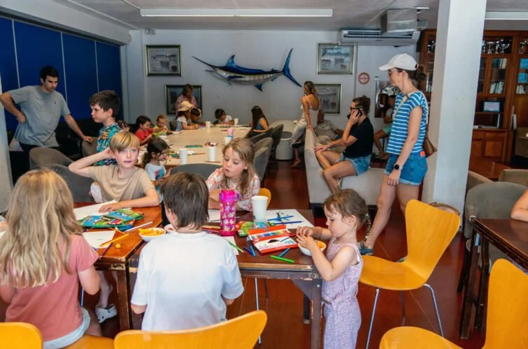 Niños y adultos en una sala, algunos niños sentados en mesas pintando y otros adultos observando, en el fondo hay un pez espada decorativo y cuadros en la pared.