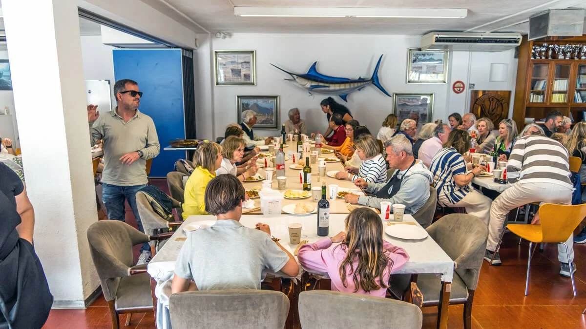 Grupo de personas en una comida o celebración en un restaurante o comedor, con un pez espada montado en la pared de fondo.