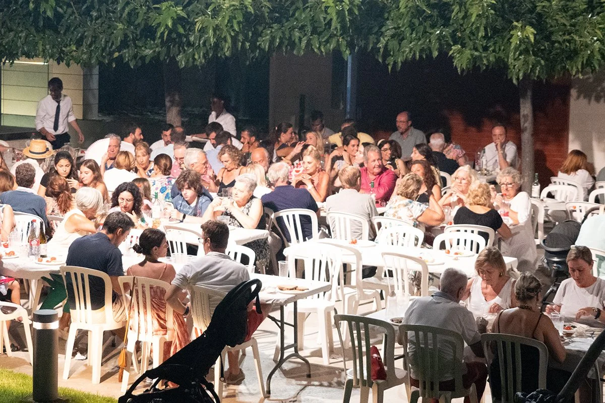 Una reunión o fiesta al aire libre con muchas personas sentadas en mesas, comiendo y conversando, bajo árboles en la noche.