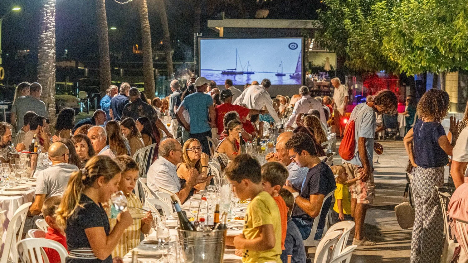 Mesa larga con muchas personas en una reunión al aire libre por la noche, con una pantalla grande en el fondo que muestra barcos sobre el agua.