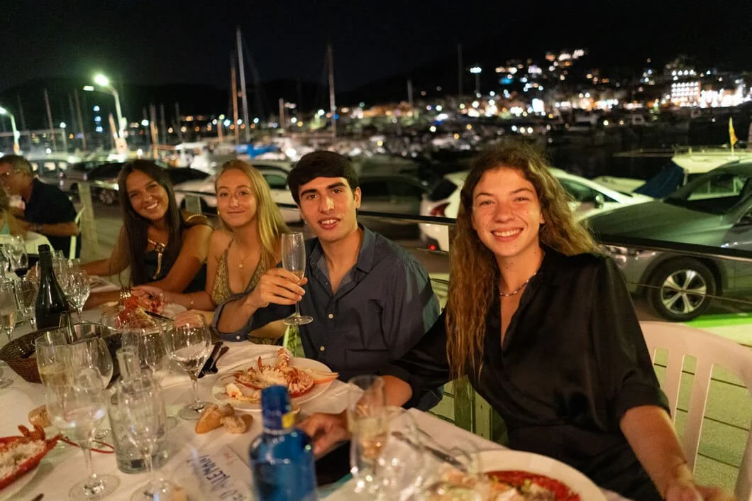 Cuatro jóvenes disfrutando una cena en un restaurante al aire libre en la noche, con un muelle y yates de fondo.
