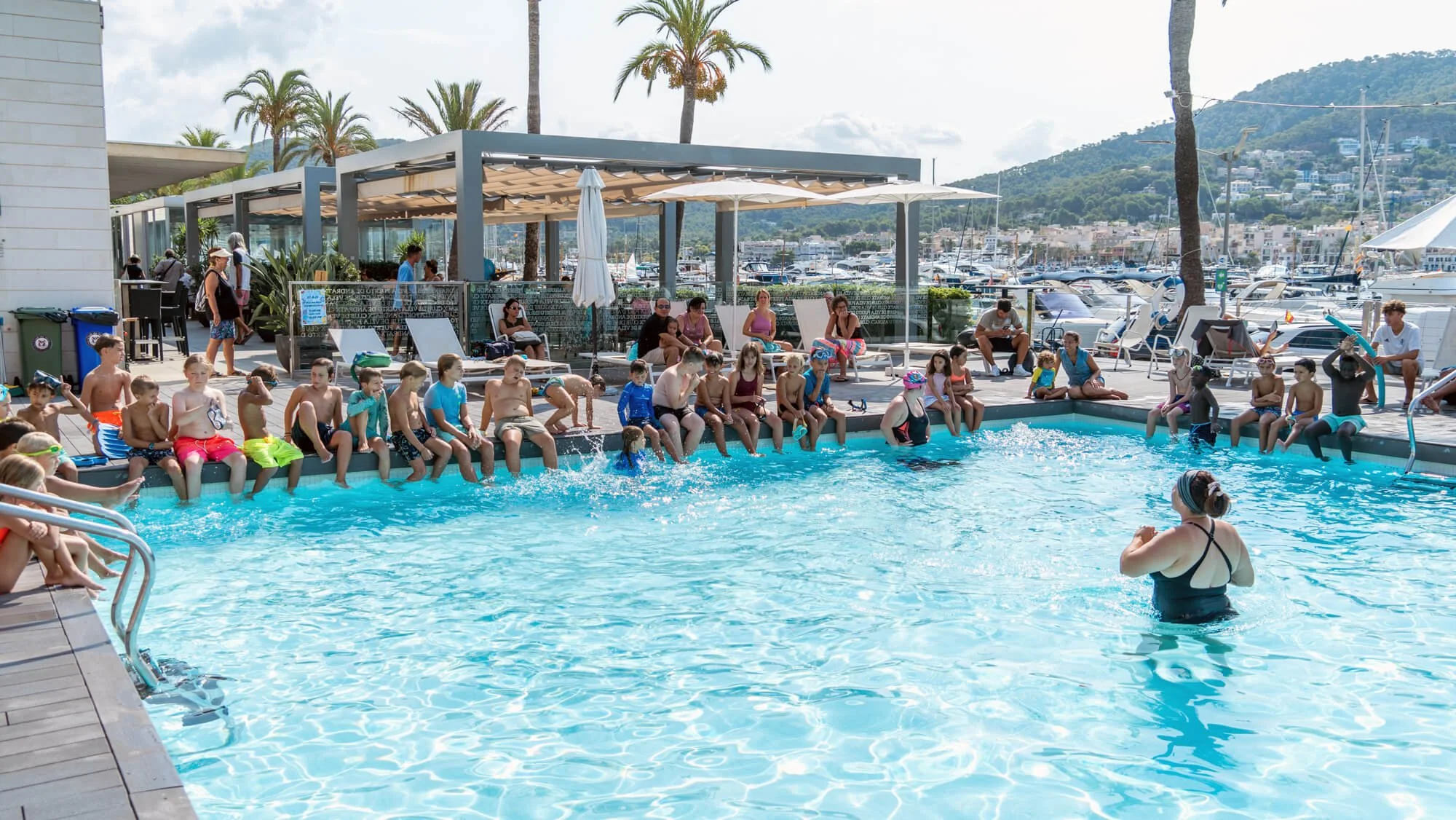 Niños y adultos disfrutando en una alberca bajo el sol, con una vista de mar y montañas al fondo, en un club o centro recreativo con sombrillas y personas sentadas en bancos y sillas alrededor.