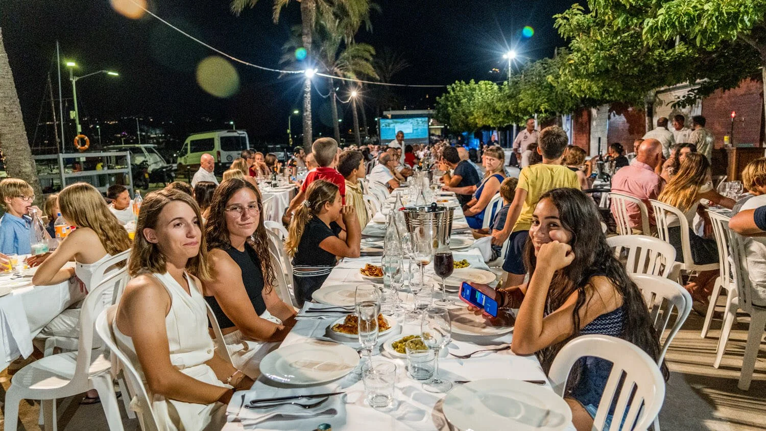Cena al aire libre en la noche con muchas personas sentadas en mesas decoradas, disfrutando de comida y bebida, en un entorno con árboles y luces. En el fondo se ven vehículos y un proyector en una pantalla.