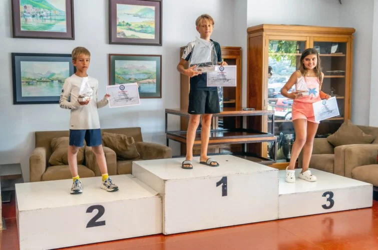 Tres niños en un podio de premiación con certificados y trofeos, en un lugar con paredes con cuadros y muebles de madera.