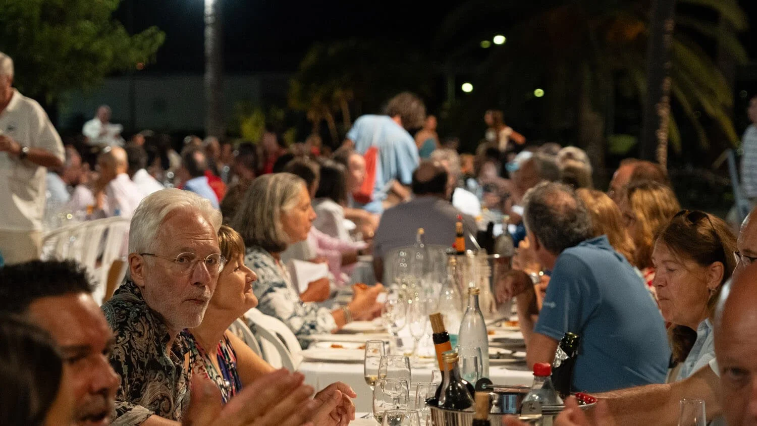Reunión de personas adultas en una comida al aire libre por la noche, sentados alrededor de una mesa con bebidas y comida.