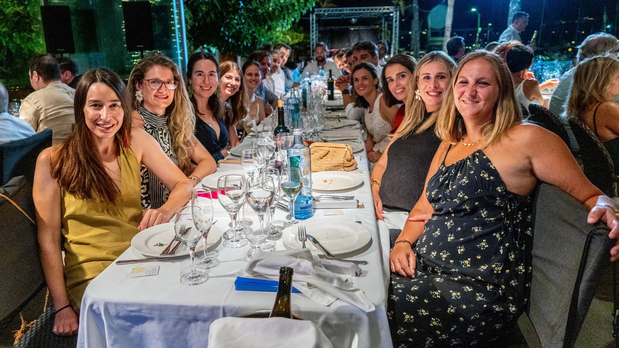Grupo de personas sentadas en una mesa en una celebración nocturna al aire libre, con alimentos y bebidas en la mesa.