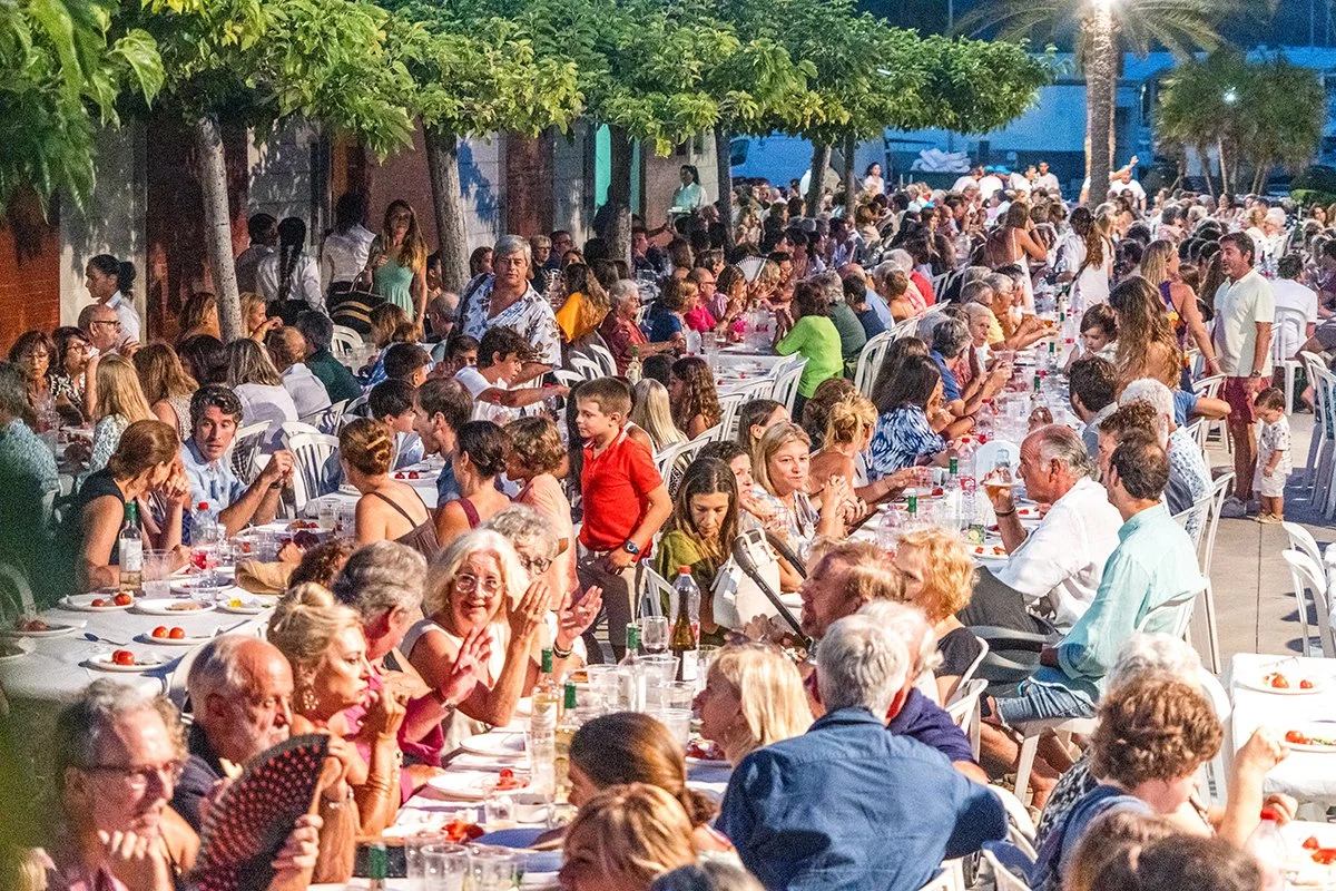 Una gran reunión al aire libre con muchas personas sentadas en mesas largas, compartiendo y disfrutando una comida en un ambiente social comunitario.