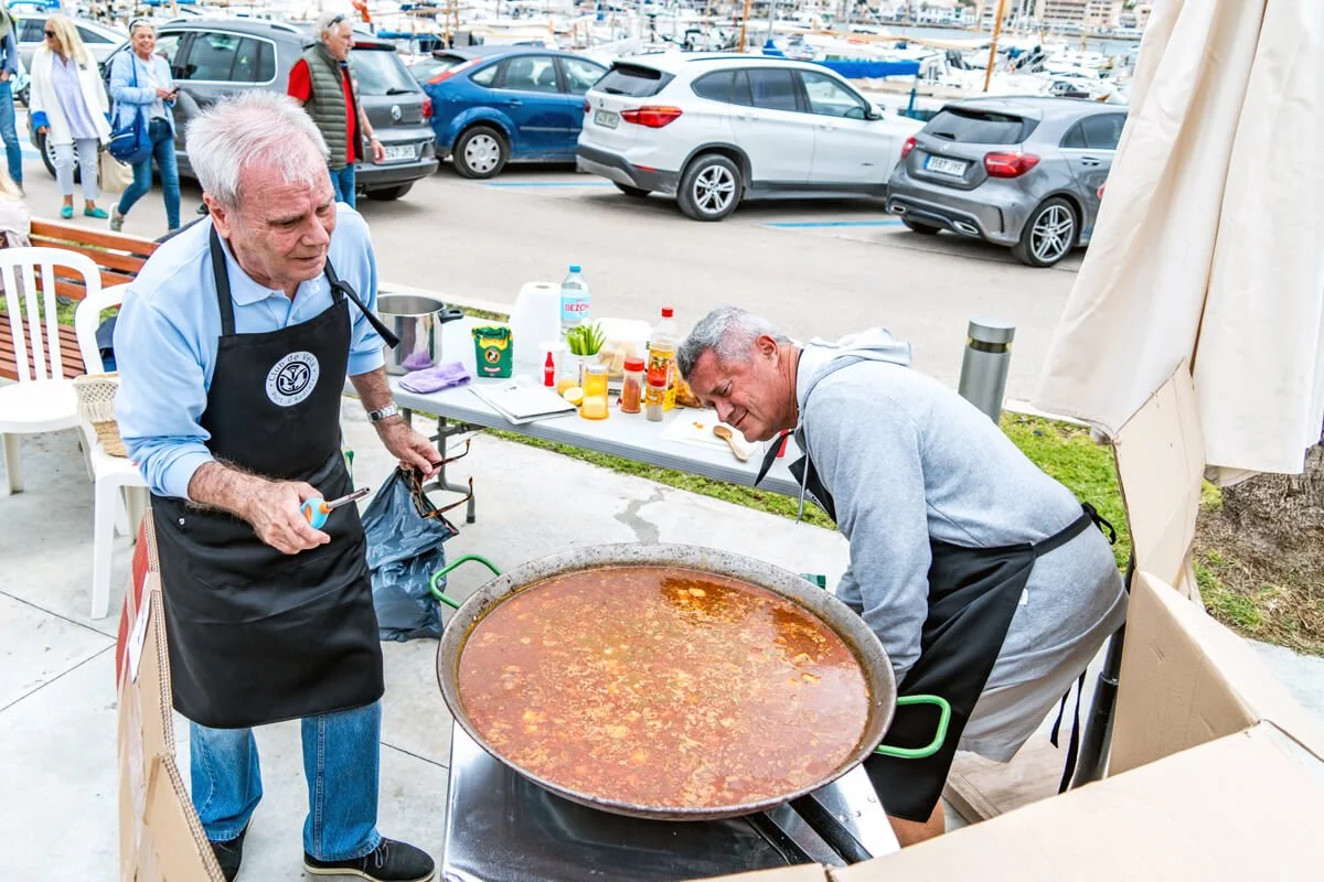 Dos hombres cocinando un caldero grande de caldo o sopa en una calle, con coches y personas en el fondo.