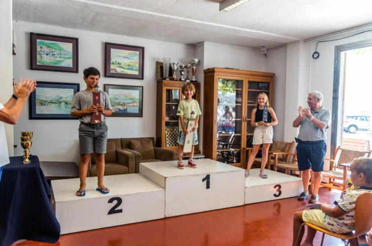 Niños en un podio de premiación recibiendo trofeos en un ambiente interior con adultos que los felicitan.